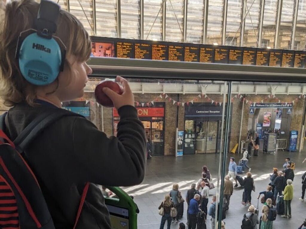 Oliver wearing blue ear defenders and holding an apple while watching the departure boards at King’s Cross Station.