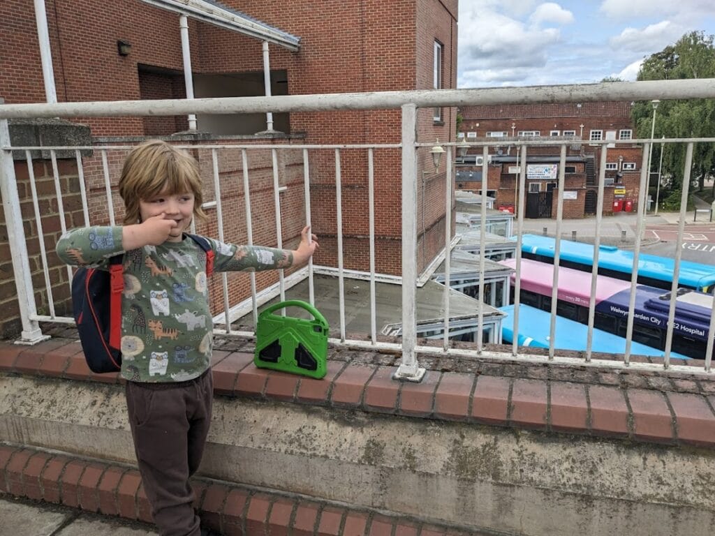 Oliver standing by a railing with his backpack on, pointing playfully while waiting near a bus station.