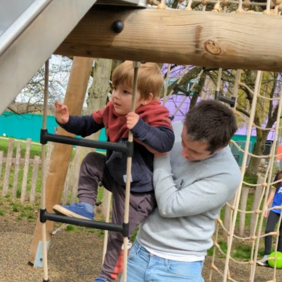 Oliver climbing a play structure with his dad supporting him from behind.