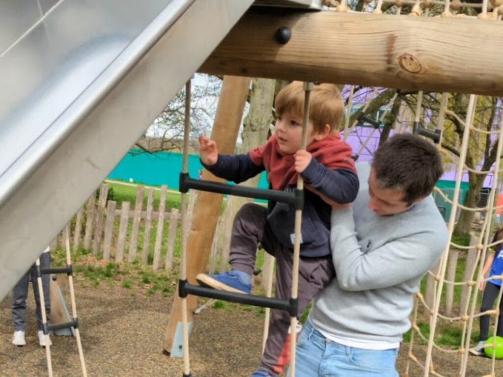 Oliver climbing a play structure with his dad supporting him from behind.